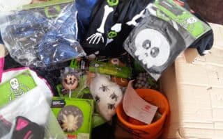 A cardboard box filled with Halloween-themed items, including skeleton costumes, a pumpkin bucket, fake spider webs with plastic spiders, chocolate treats, and a black bag with a white skull design. Packaging and a shopping receipt are also visible.