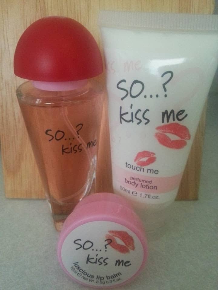 Three "So...? Kiss Me" products: a perfume bottle with a red cap, a white body lotion tube labelled "touch me," and a round tub of luscious lip balm, all decorated with red lipstick kiss prints, are displayed upright on a light surface.