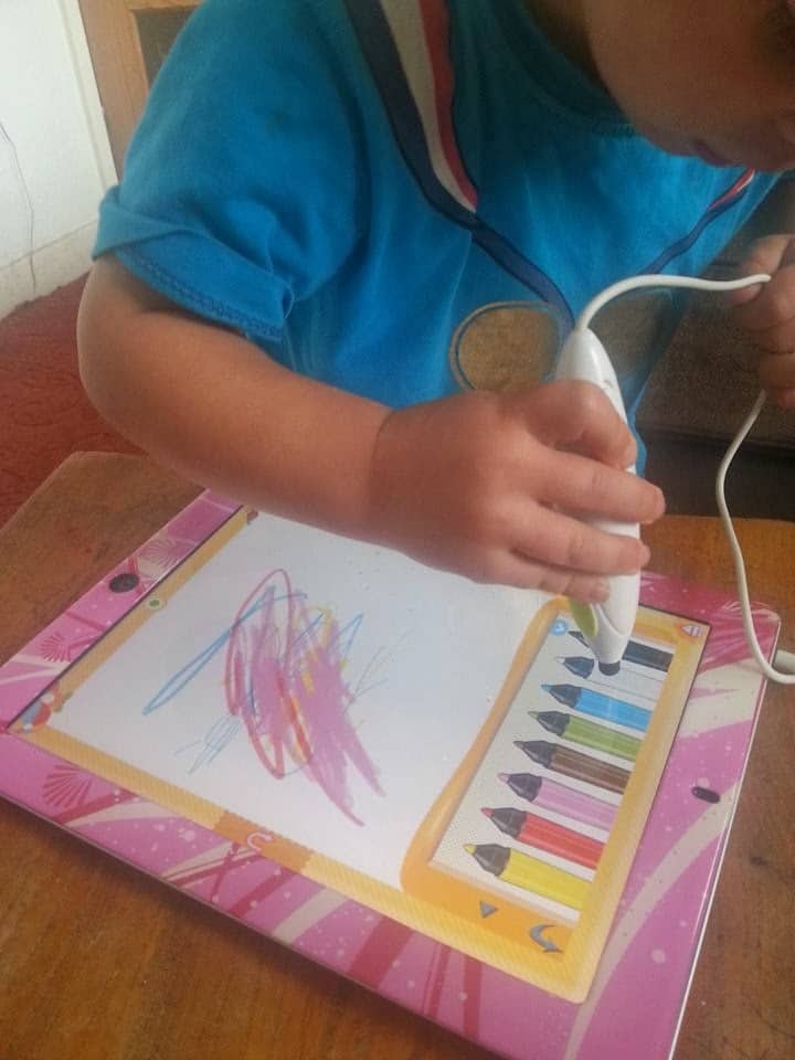 A young child in a blue shirt uses a white electric toothbrush to colour on a drawing pad, making colourful scribbles with the brush instead of crayons. The drawing pad has cartoon crayon designs on its border and sits on a wooden table.