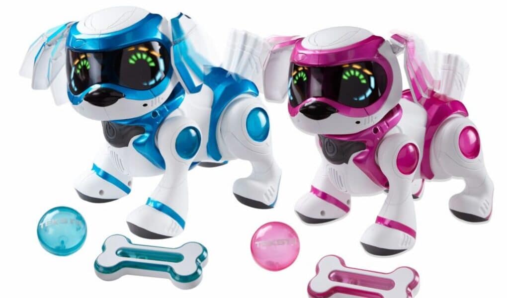 Two robotic toy dogs, one blue and one pink, each with large black visors displaying animated eyes. They stand beside matching coloured bones and balls. The dogs have white bodies with coloured accents, futuristic features, and appear playful. The background is plain white.