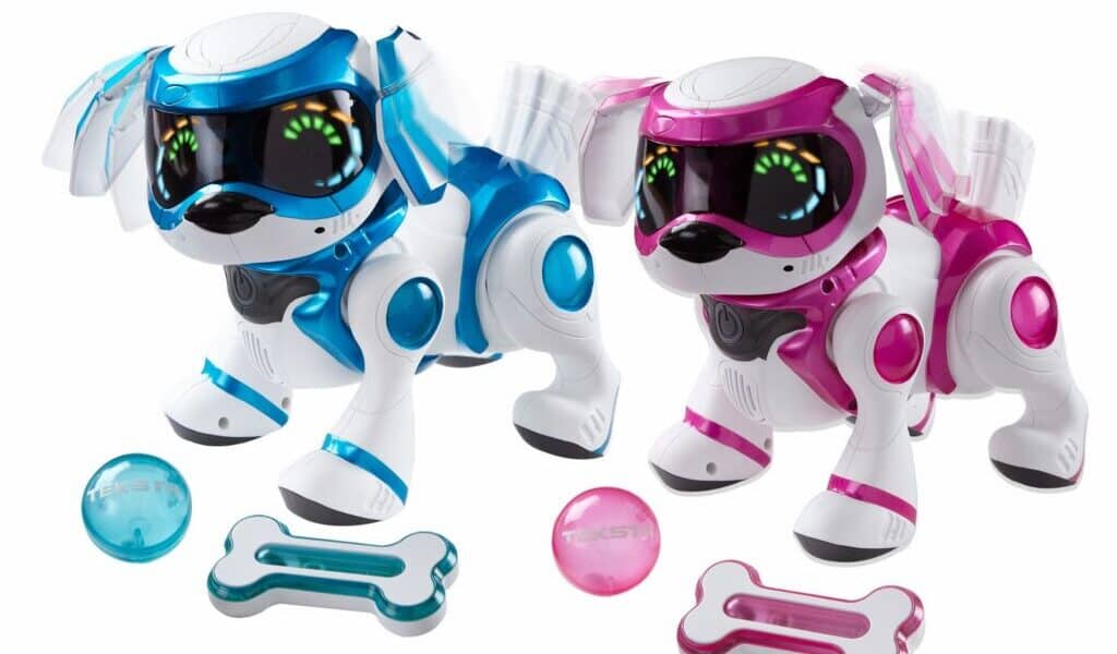 Two robotic toy dogs, one blue and one pink, each with large black visors displaying animated eyes. They stand beside matching coloured bones and balls. The dogs have white bodies with coloured accents, futuristic features, and appear playful. The background is plain white.