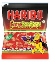 A bag of HARIBO Fangtastics sweets features a red, black, and green design. The yellow HARIBO bear mascot, dressed as a vampire, holds a bat. The bag displays red and white fang-shaped jelly sweets through a transparent window.