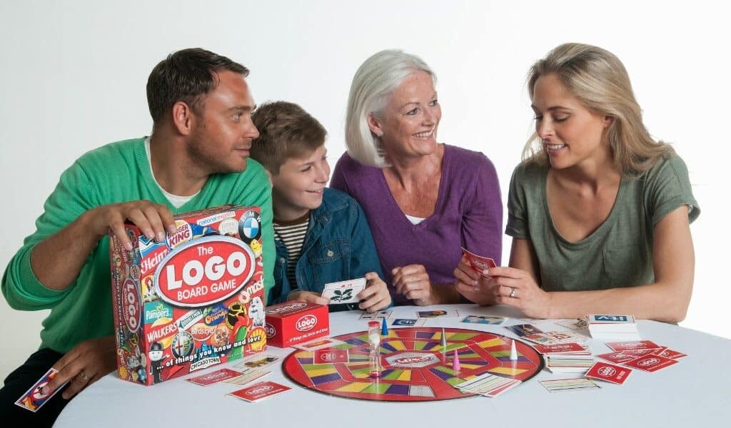 Four people—two adults, a senior woman, and a child—sit around a round table playing the Logo Board Game. They are smiling, holding cards, and interacting with the colourful game board and pieces on the white table. The background is plain white.
