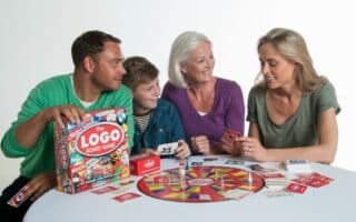 Four people—two adults, a senior woman, and a child—sit around a round table playing the Logo Board Game. They are smiling, holding cards, and interacting with the colourful game board and pieces on the white table. The background is plain white.