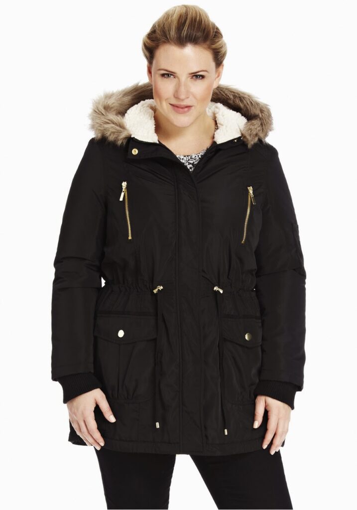 A woman stands facing forwards, wearing a black parka coat with a faux fur-lined hood, gold zips, and buttoned pockets. She has fair skin, blonde hair pulled back, and is smiling slightly. The background is plain white.