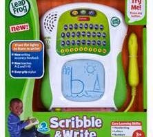 A LeapFrog Scribble & Write toy in its packaging. The device has a green and white body with a letter keypad, speaker, and a blue drawing screen showing childlike writing. A yellow stylus is attached. The box features a smiling child and colourful product details.