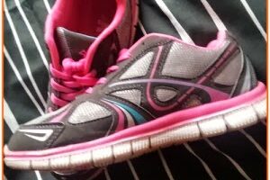 A pair of black and grey trainers with pink accents and white soles rests on a black fabric with white stripes. One trainer is upright, while the other lies on its side, revealing the pink interior and laces.