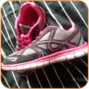 A pair of black and grey trainers with pink accents and white soles rests on a black fabric with white stripes. One trainer is upright, while the other lies on its side, revealing the pink interior and laces.