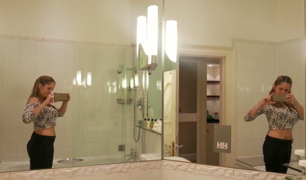 A woman stands in a bathroom taking a mirror selfie with her phone. She wears a long-sleeved, fitted, patterned crop top and dark trousers. The reflection shows both her front and back. The bathroom is well-lit with white tiles, a bath, and shelves in the background.