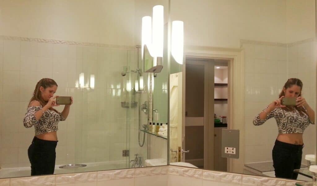 A woman stands in a bathroom taking a mirror selfie with her phone. She wears a long-sleeved, fitted, patterned crop top and dark trousers. The reflection shows both her front and back. The bathroom is well-lit with white tiles, a bath, and shelves in the background.