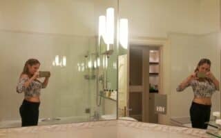 A woman stands in a bathroom taking a mirror selfie with her phone. She wears a long-sleeved, fitted, patterned crop top and dark trousers. The reflection shows both her front and back. The bathroom is well-lit with white tiles, a bath, and shelves in the background.
