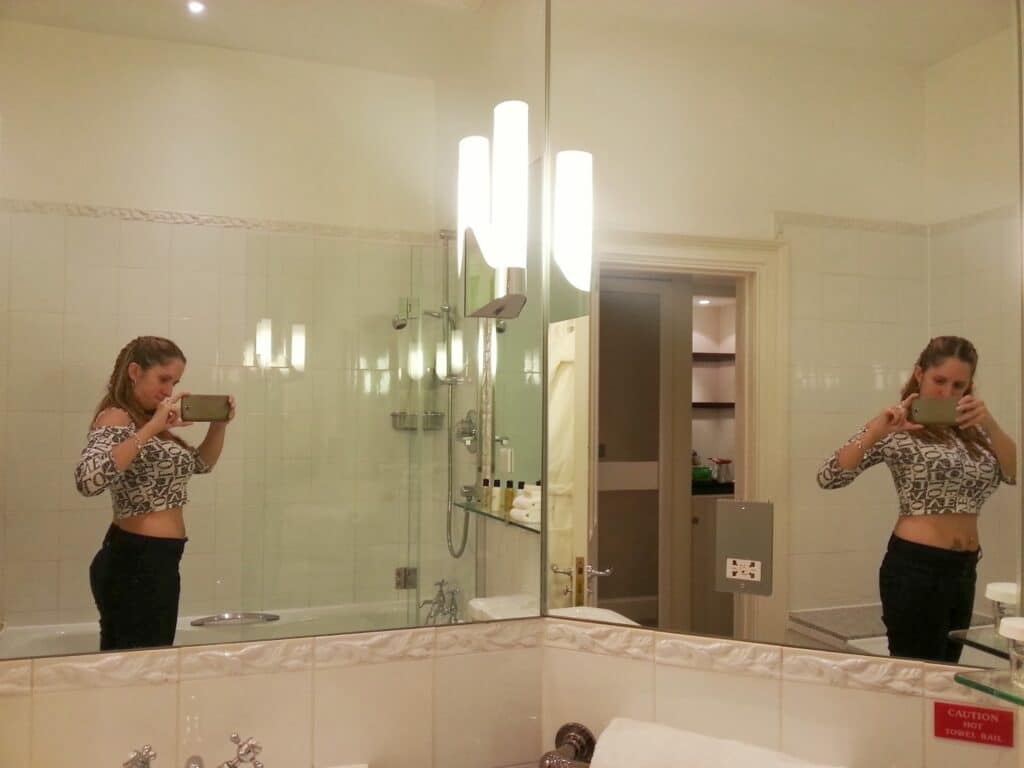 A woman in a crop top and dark trousers takes a mirror selfie in a well-lit bathroom. The large mirror reflects her side and front view. The bathroom has a bath, white tiles, and shelves visible in the background.