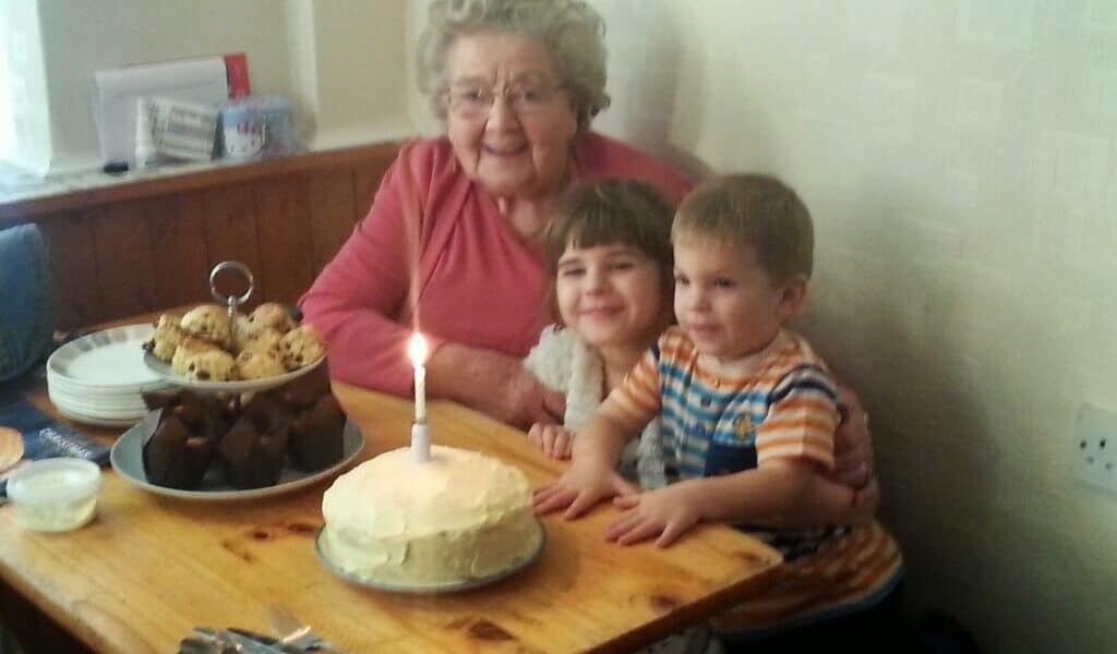 An elderly woman sits at a wooden table with two young children. In front of them is a cake with a single lit candle, as well as a plate of brownies and fairy cakes. They are all smiling, appearing to celebrate a birthday in a cosy room.