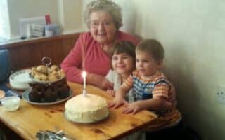 An elderly woman sits at a wooden table with two young children. In front of them is a cake with a single lit candle, as well as a plate of brownies and fairy cakes. They are all smiling, appearing to celebrate a birthday in a cosy room.
