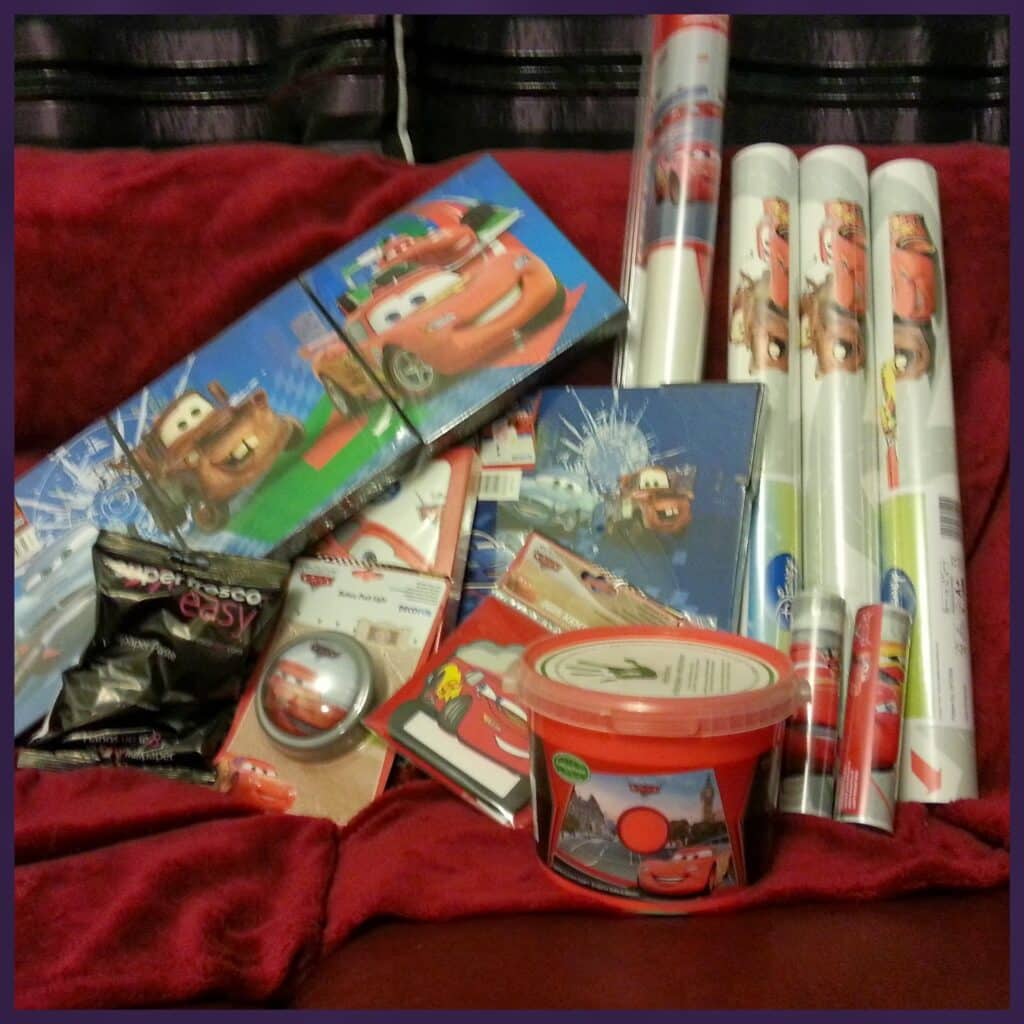 A collection of Disney Pixar Cars-themed items sits on a red blanket, including wrapping paper rolls, notebooks, a gift bag, greeting cards, a bucket, a spoon, and stickers, mostly featuring Lightning McQueen and Mater characters.