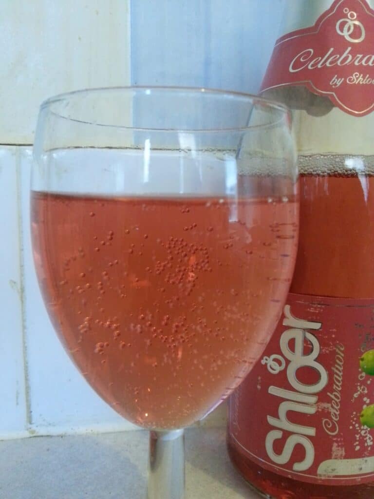 A glass of sparkling pink drink with bubbles sits in front of a bottle labelled “Shloer Celebration.” Both the drink and bottle are pink. The background features white tiles and a light-coloured countertop.