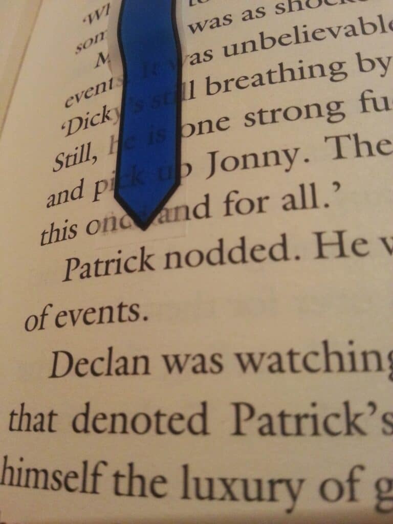 A close-up of an open book page with a blue arrow-shaped bookmark pointing downwards. The visible text includes the sentence, “Patrick nodded. He was…” and parts of other sentences. The background is softly lit and focused on the bookmark.