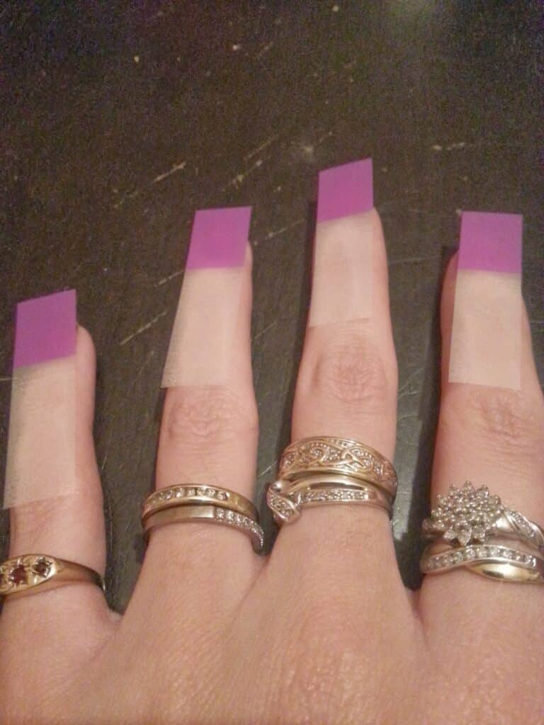 A hand with four fingers visible, each wearing ornate gold rings. Each finger has a long, rectangular false nail attachment with a clear base and a purple tip, creating the effect of unusually elongated, square-shaped nails. The background is dark.