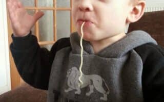 A young boy with fair hair, wearing a grey and black Lonsdale London sweatshirt, stands indoors with arms raised. He has a single long noodle hanging from his mouth, looking playful and slightly smug. A glass door and brown sofa are in the background.