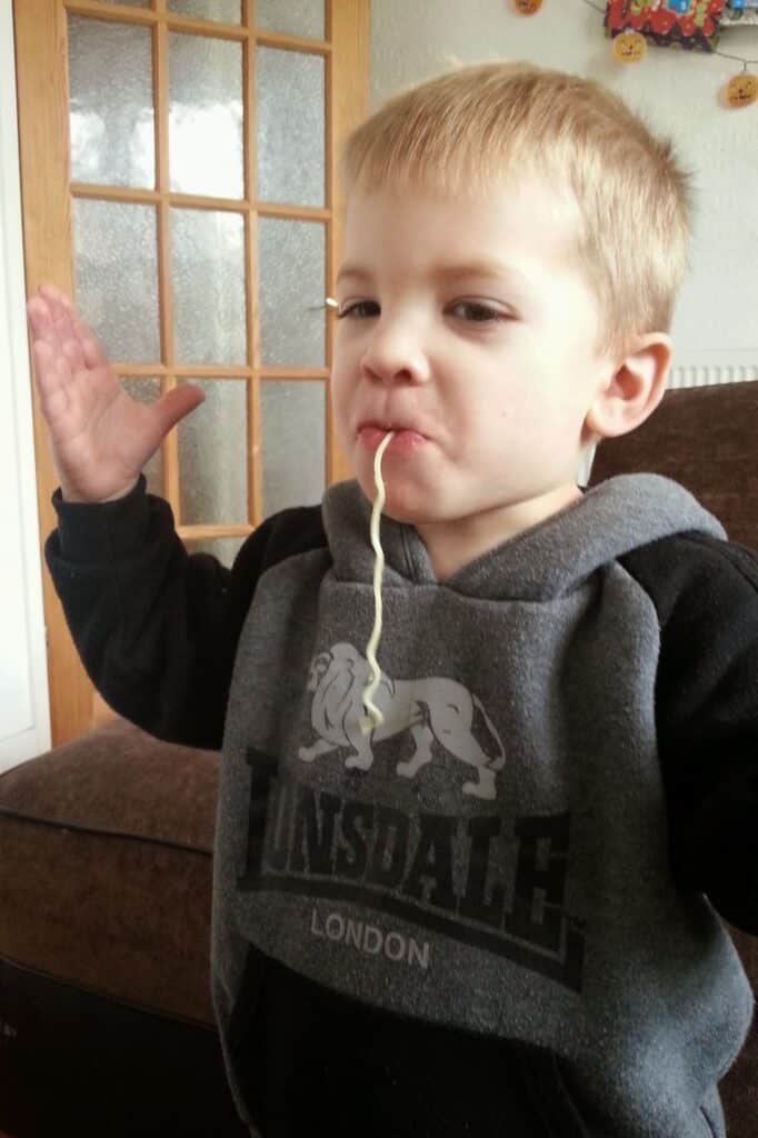 A young boy in a grey Lonsdale London hoodie stands indoors with one hand raised and a strand of spaghetti hanging from his mouth. He looks slightly amused and confident. In the background, there is a brown sofa and a glass-panelled door.