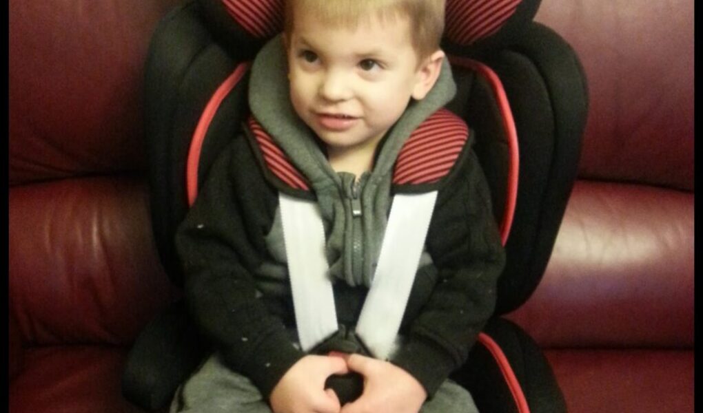 A young child with short light brown hair sits in a black and red car seat placed on a maroon sofa. The child is wearing a grey hoodie and grey tracksuit bottoms, buckled securely, and looks slightly to the side with a small smile.