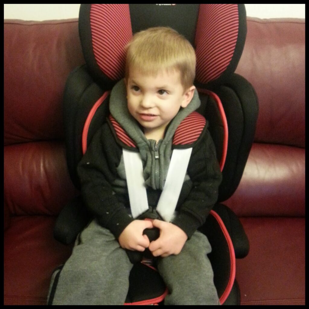A young child with short, light brown hair sits in a black and red car seat placed on a maroon sofa. The child is wearing a grey outfit with a black hoodie and is looking slightly to the side with a small smile.