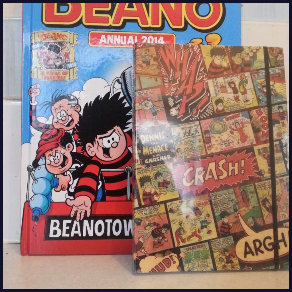 Two comic books stand upright: one is “BEANO Annual 2014” featuring cartoon characters including Dennis the Menace and Gnasher on a blue, red, and yellow cover; the other has a collage of vintage comic panels with bold words like “CRASH!” and “ARGH!”.