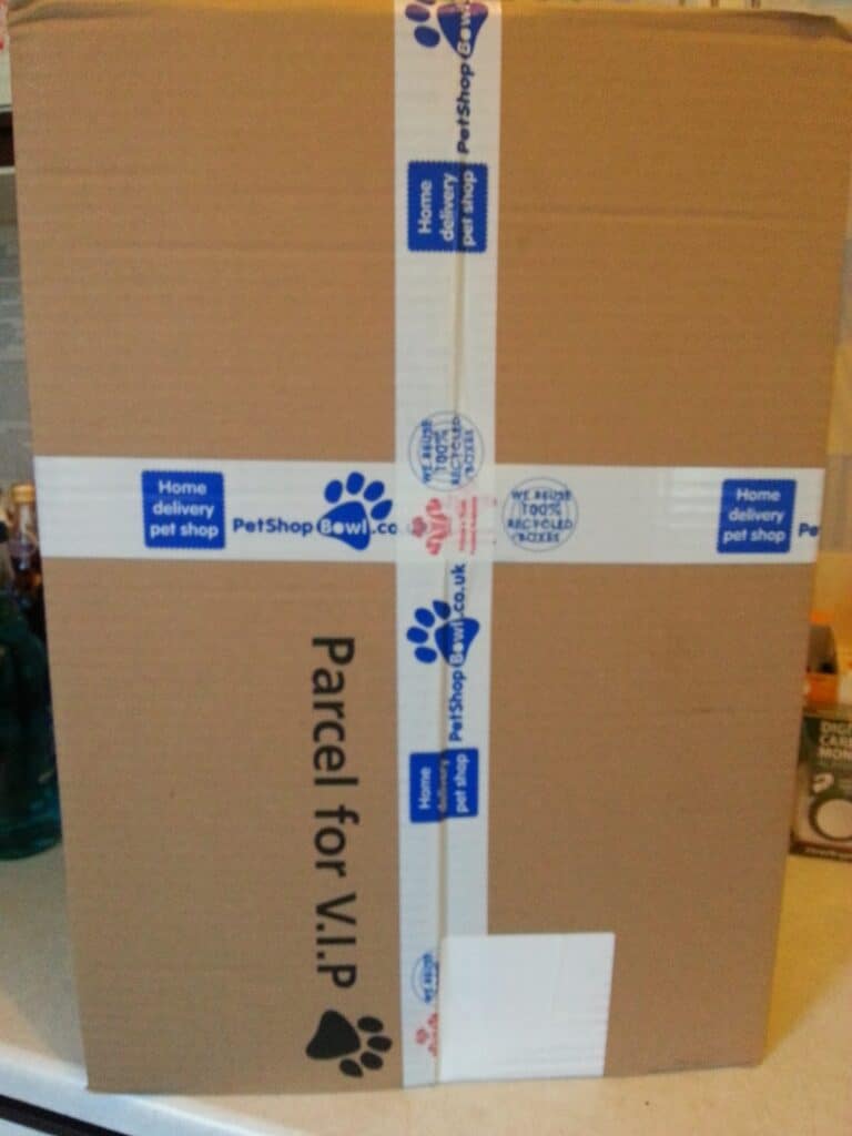 A large cardboard box labelled “Parcel for V.I.P” stands upright. The box is sealed with white tape decorated with blue paw prints and “PetShopBowl.co.uk Home delivery pet shop” branding. The background shows shelves with various household items.