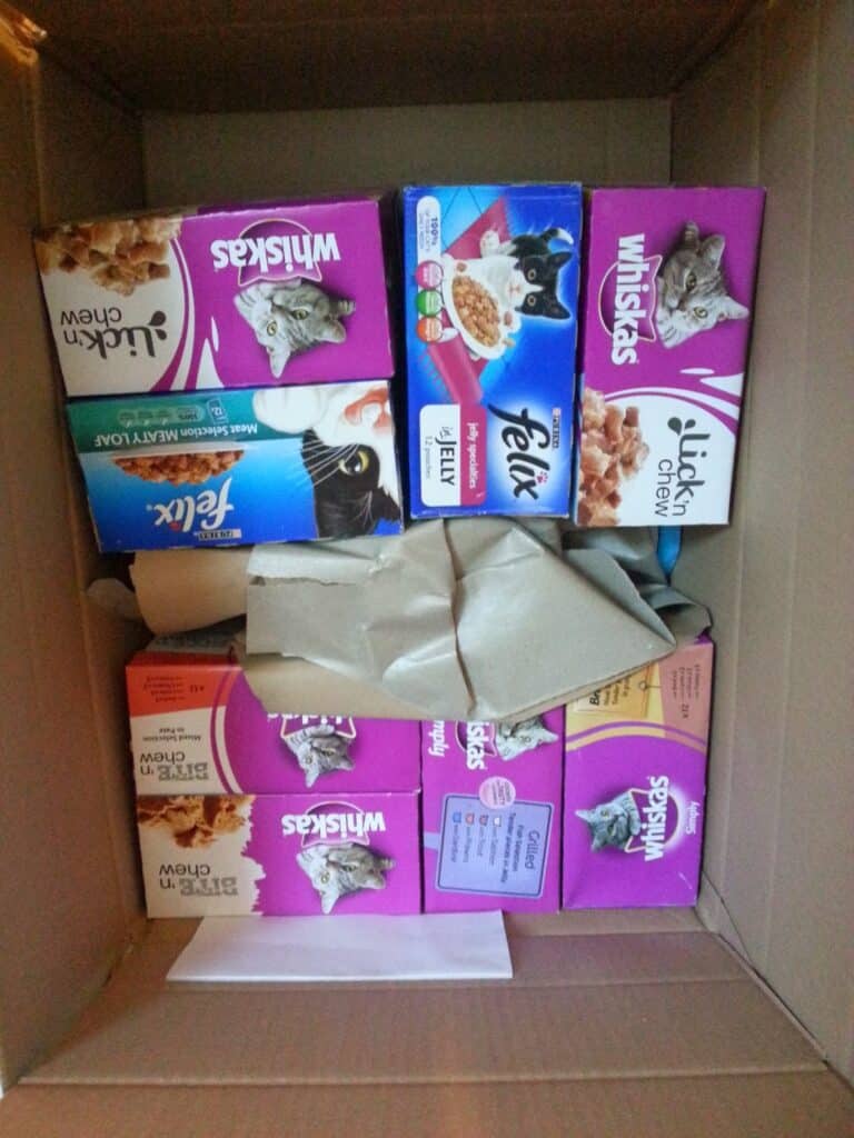 A cardboard box filled with various boxes of cat food, including Whiskas and Felix brands, in purple, blue, and orange packaging. Some boxes show pictures of cats, and there is brown packing paper tucked among the boxes.