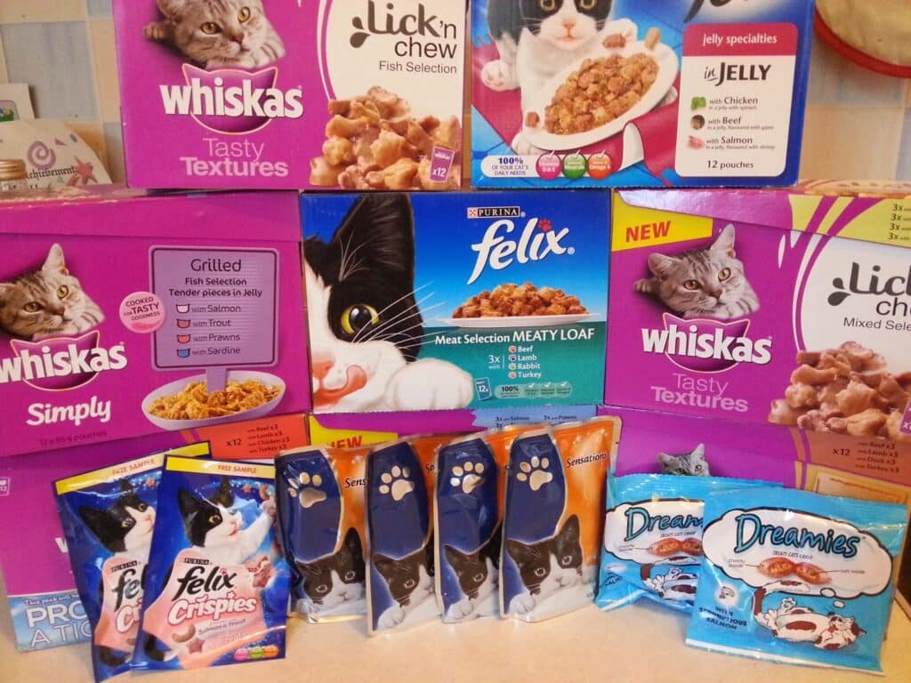 An assortment of cat foods and treats is displayed. Brands include Whiskas, Felix, Lick'n'Chew, and Dreamies. Colourful boxes and packets feature images of cats and food, with a variety of flavours and textures shown on the packaging.