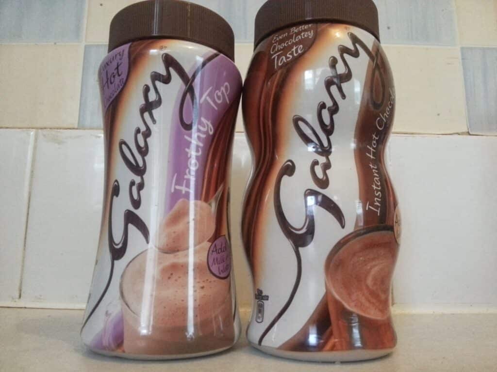 Two jars of Galaxy instant hot chocolate powders stand side by side on a counter. The left jar is labelled "Instantly Top" with a light purple label and creamy drink image; the right jar says "Instant Hot Chocolate" with a brown label and a frothy chocolate drink image.