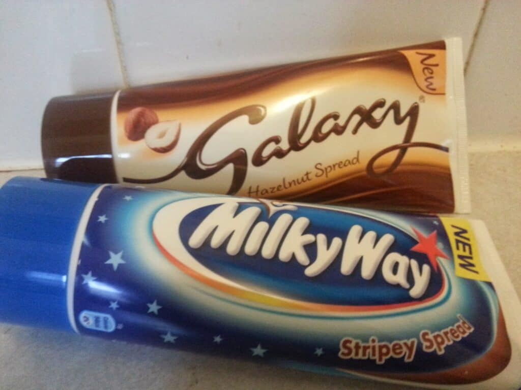 Two tubes of spread are shown: one is Galaxy Hazelnut Spread with a hazelnut image and brown-gold swirl design, the other is Milky Way Stripey Spread with a blue starry design. Both have "NEW" labels on the top right corners.