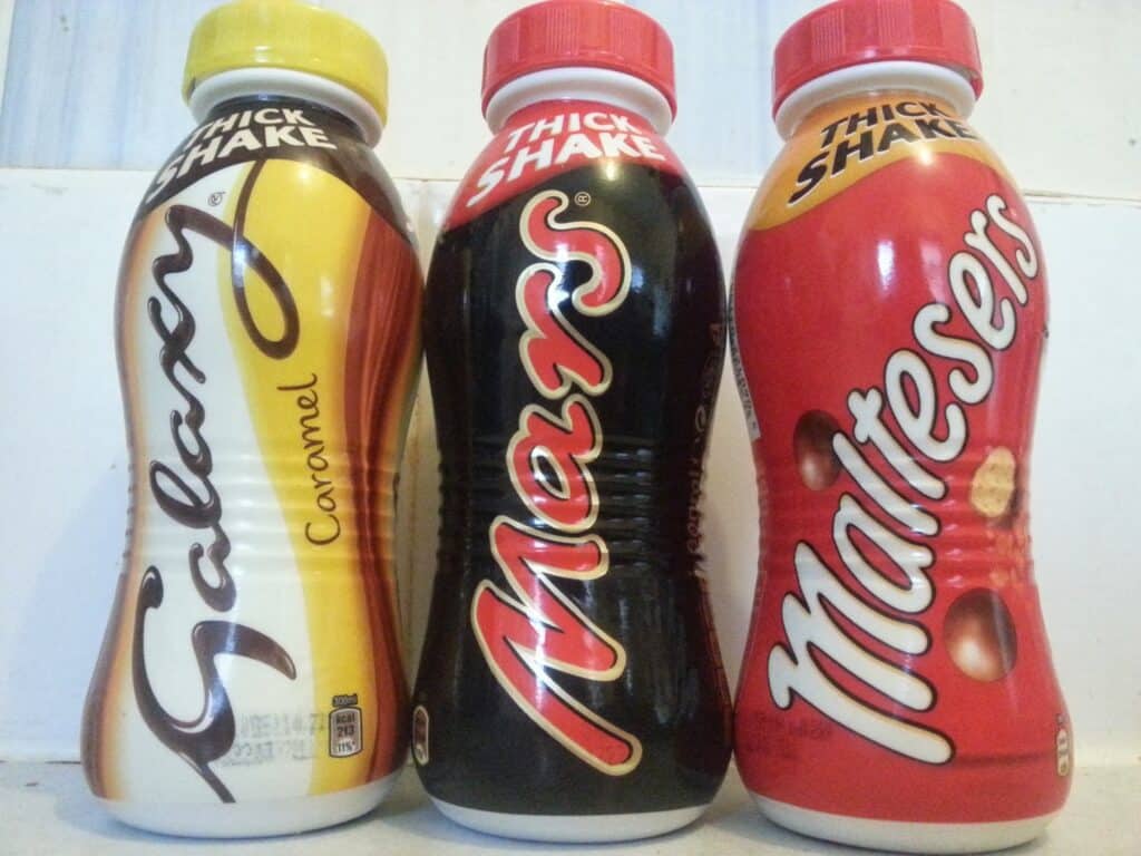 Three thick shake bottles stand side by side: Galaxy Caramel (yellow and brown), Mars (black and red), and Maltesers (red with malt balls). Each bottle features a white cap and brand logo prominently on the front.