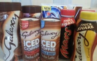Assorted chocolate drinks on a counter, including Galaxy Iced Coffee (Mocha Latte Double Shot, Mocha Latte Single Shot), Galaxy Thick Shake Caramel, Galaxy drinking chocolate, Mars Thick Shake, and a partially visible Milky Way product in the back.