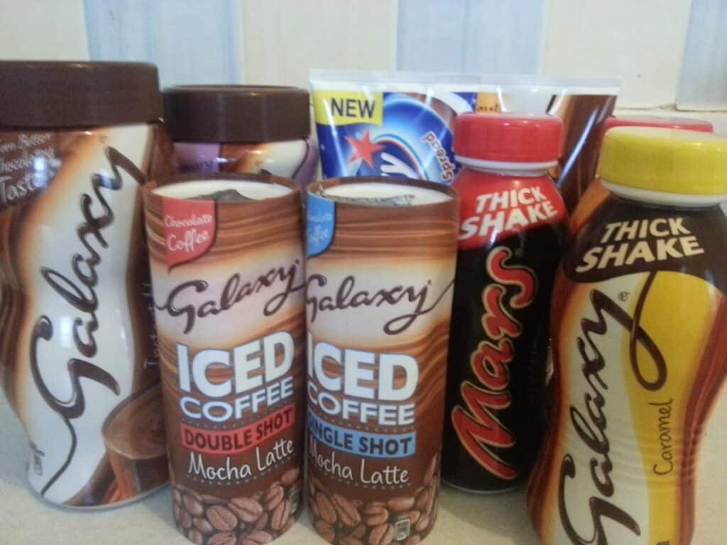 A group of chilled drink bottles and tubs is arranged together, including Galaxy Iced Coffee in Mocha Latte and Single Shot flavours, Mars Thick Shake, Galaxy Thick Shake Caramel, and a container of Nestlé Milkybar and Smarties ice cream. Labels are clearly visible.