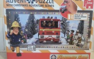 Box for a “Fireman Sam” advent puzzle. The cover shows Fireman Sam running, a fire engine, children, and numbered doors in a snowy, festive scene with Christmas lights. Text says “24 puzzle,” “With Christmas adventure scenes,” and “Open up an adventure window each day.”.
