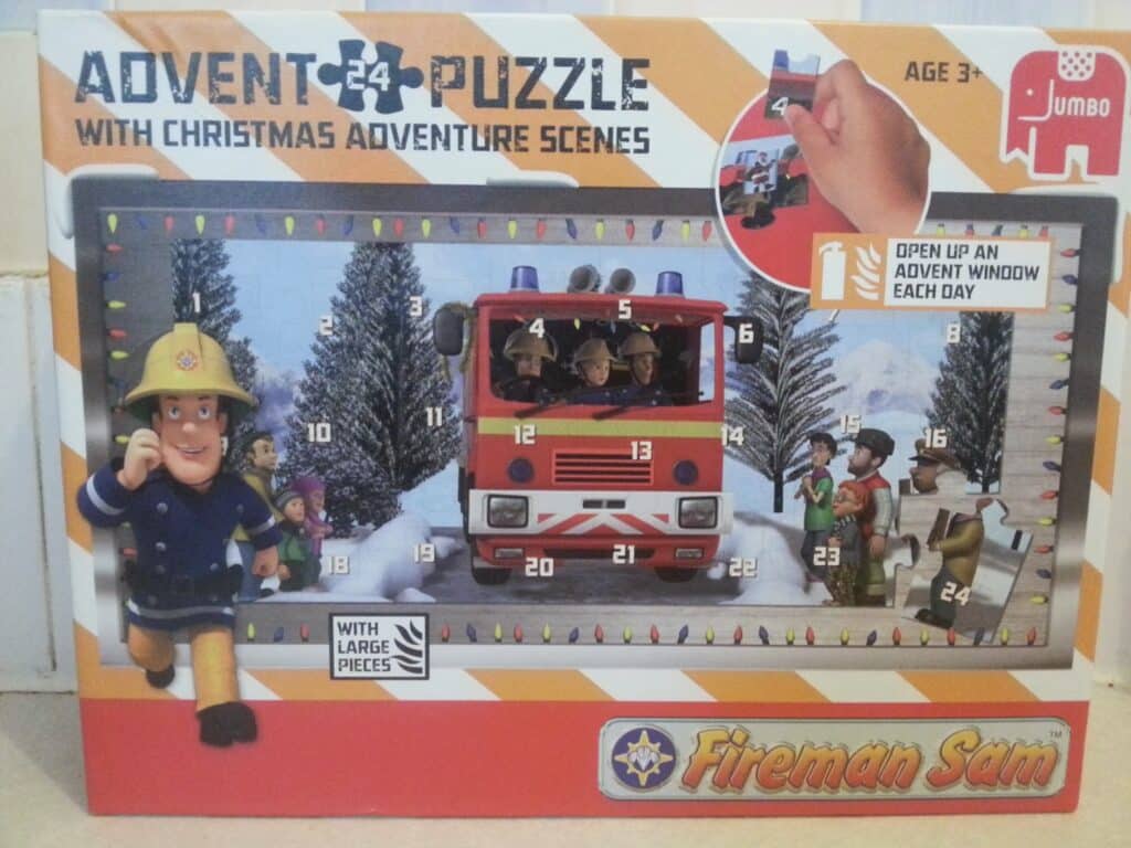 A Fireman Sam Advent Puzzle box features a festive scene with Fireman Sam beside a decorated fire engine. Numbered windows 1–24 cover the scene, surrounded by snowy trees and Christmas lights. The box promotes “Christmas adventure scenes” and “large pieces”.