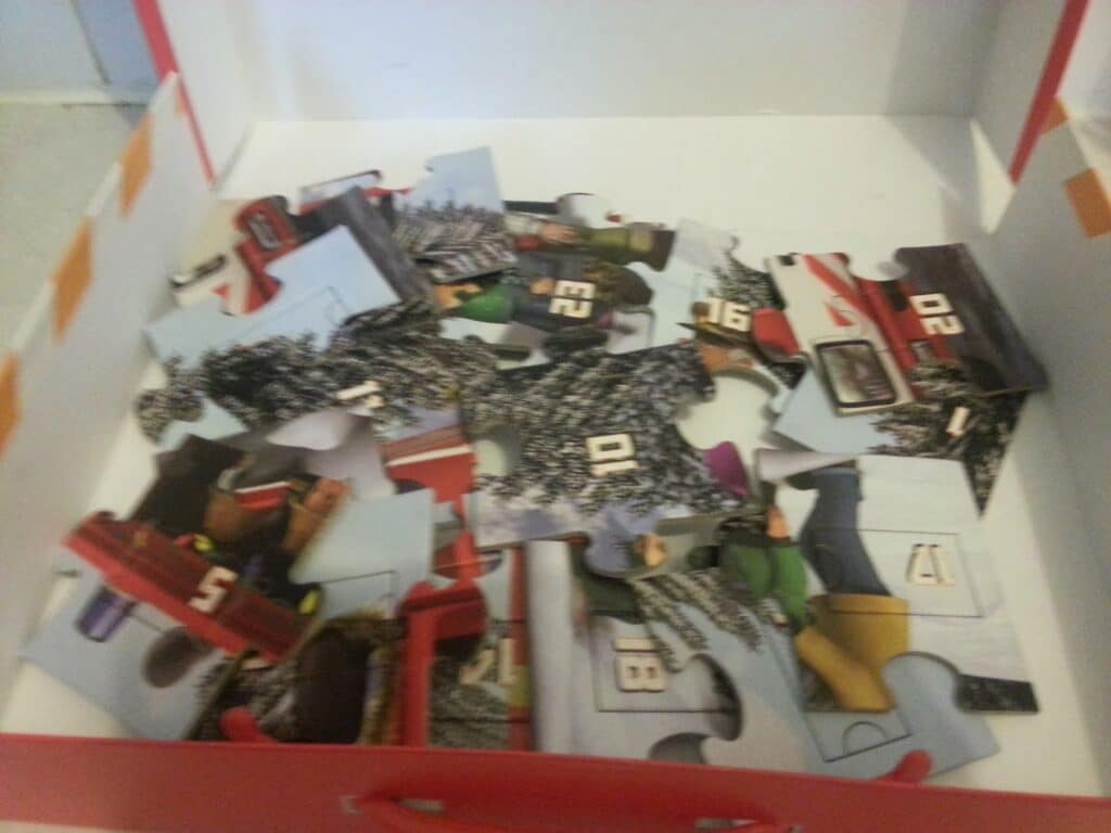 A close-up of a box containing several large, scattered jigsaw puzzle pieces with numbers, vehicles, and outdoor scenery partially visible on them. The box has a white base and red sides, and the puzzle appears to be unfinished.