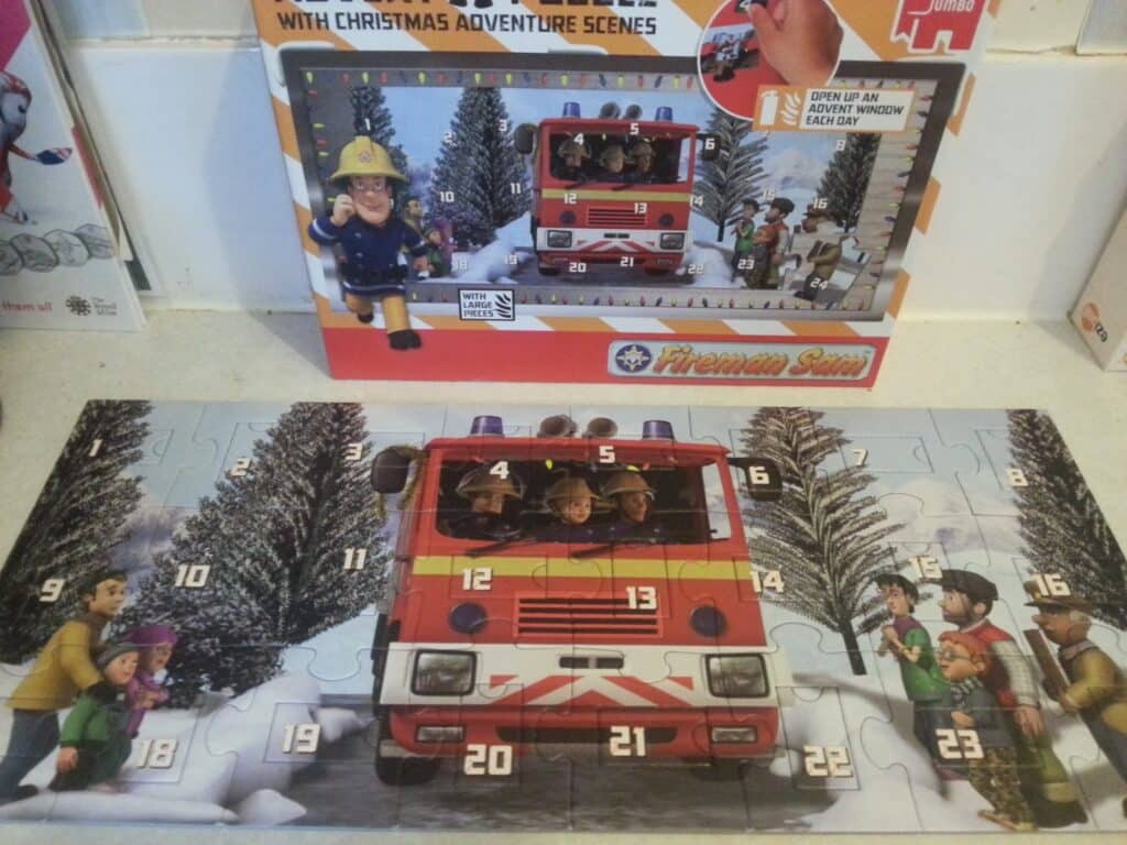 A "Fireman Sam" Christmas advent jigsaw is assembled on a table, showing characters in and around a red fire engine numbered 1–24. The puzzle box behind it displays the same image, with snowy trees and children watching beside the engine.