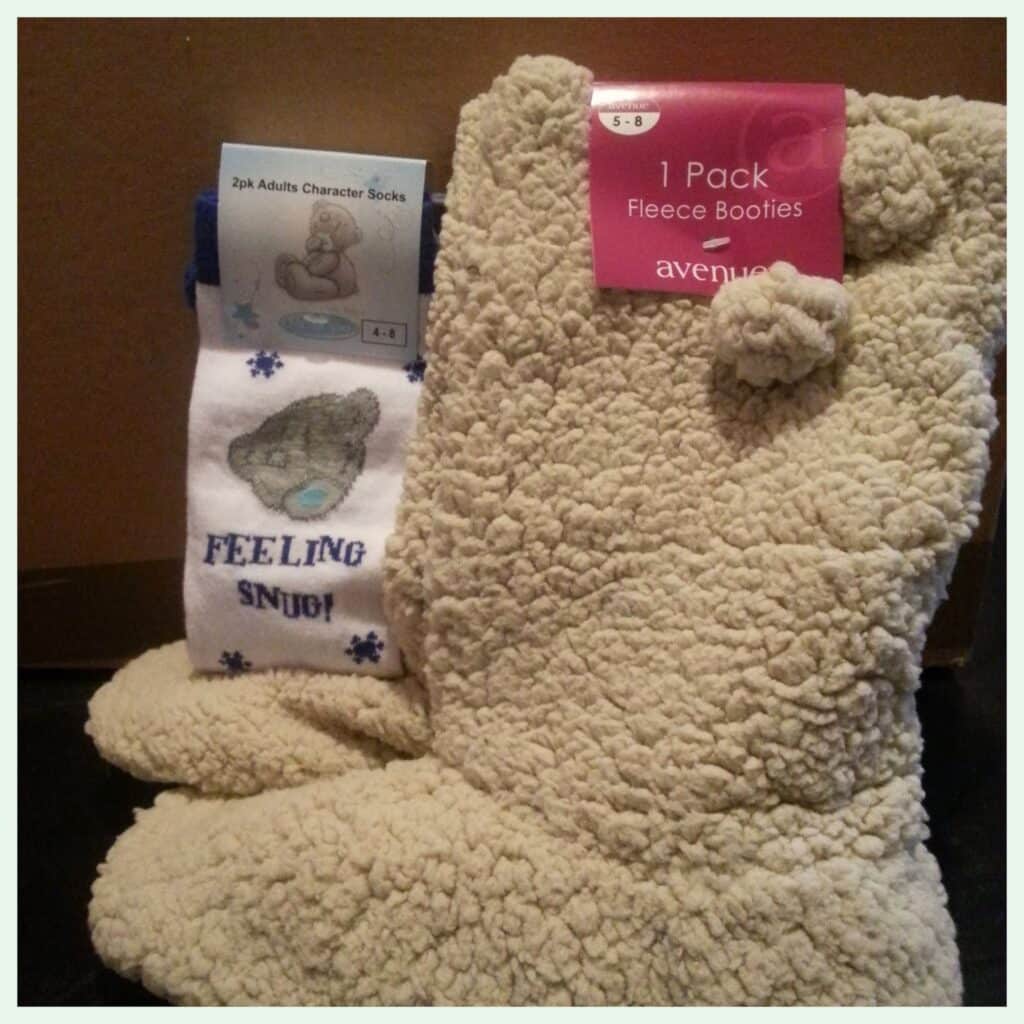 A pair of cream-coloured, fluffy fleece booties with pom-poms and a pair of white socks featuring a grey bear cartoon and blue text that says “FEELING SNUG!” Both items are placed upright against a brown background.