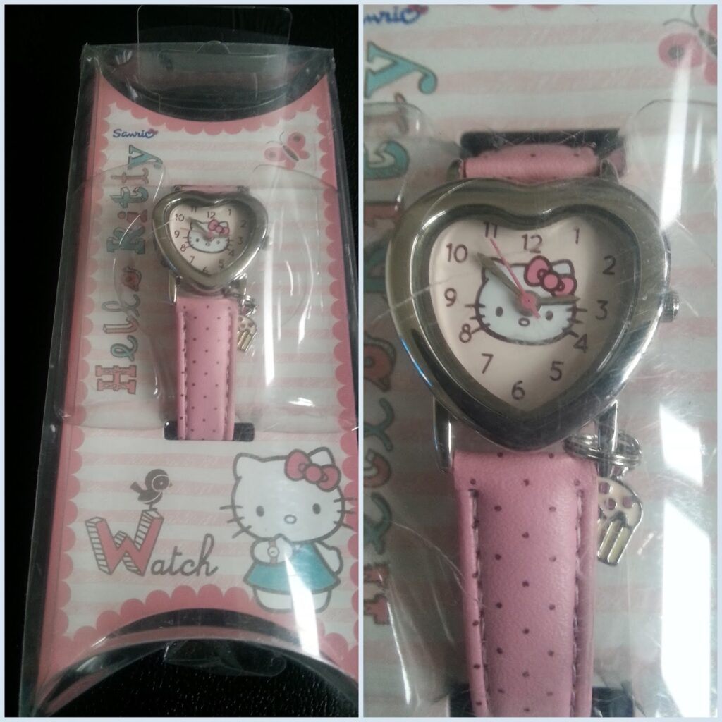 A heart-shaped Hello Kitty watch in its packaging, featuring a pink polka-dot strap and a silver case. The clock face displays Hello Kitty’s face with a pink bow. The packaging is pink with Hello Kitty graphics and the words “Hello Kitty Watch”.
