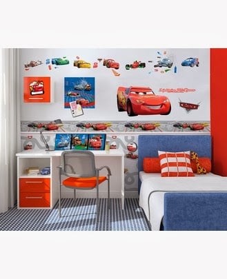 A children’s bedroom decorated with Disney Cars theme: Cars character stickers on the wall, a white desk with red drawers, an orange chair, toy cars, and a blue-and-white bed with orange, white, and chequered pillows. The floor has a blue chequered pattern.