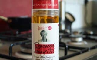 A 200ml bottle of Fussels Extra Virgin Rapeseed Spray Oil stands on a kitchen worktop in front of a stainless steel gas cooker. The bottle has a red and white label with text and a small illustration of a chef. The background is slightly blurred.