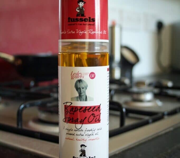 A 200ml bottle of Fussels Extra Virgin Rapeseed Spray Oil stands on a kitchen worktop in front of a stainless steel gas cooker. The bottle has a red and white label with text and a small illustration of a chef. The background is slightly blurred.