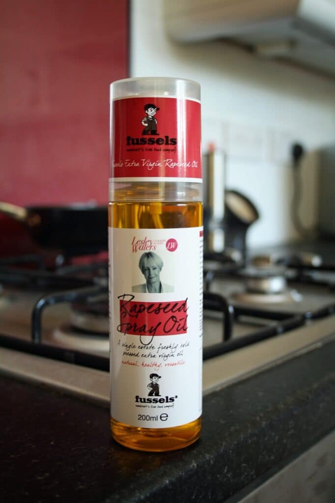 A bottle of Fussels Rapeseed Spray Oil stands on a kitchen worktop beside a gas cooker. The bottle has a red and white label with a chef’s portrait and text. The background shows a saucepan on the cooker and out-of-focus kitchen items.