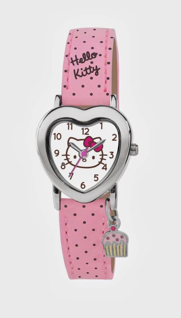 A pink polka-dot watch with a heart-shaped face featuring Hello Kitty’s face. The strap says “Hello Kitty.” A small silver cupcake charm hangs from the watch. The hour and minute hands are pink; numbers are in a playful font.