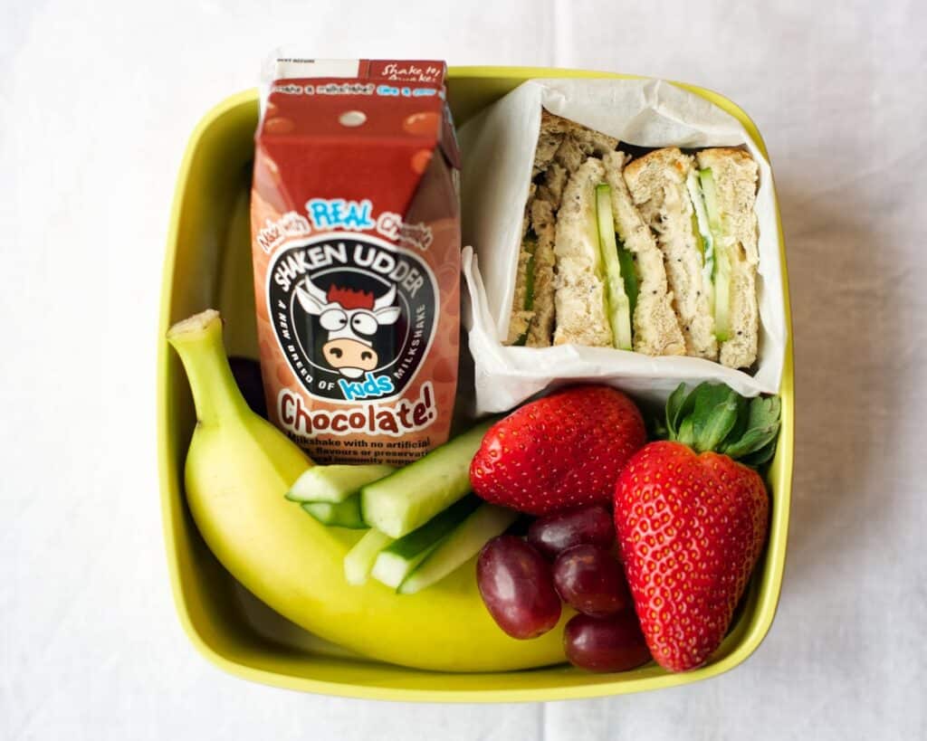A yellow lunchbox contains a chocolate milk carton, a sandwich with cucumber slices, a banana, two strawberries, a few grapes, and slices of cucumber, all arranged neatly on a white tablecloth.