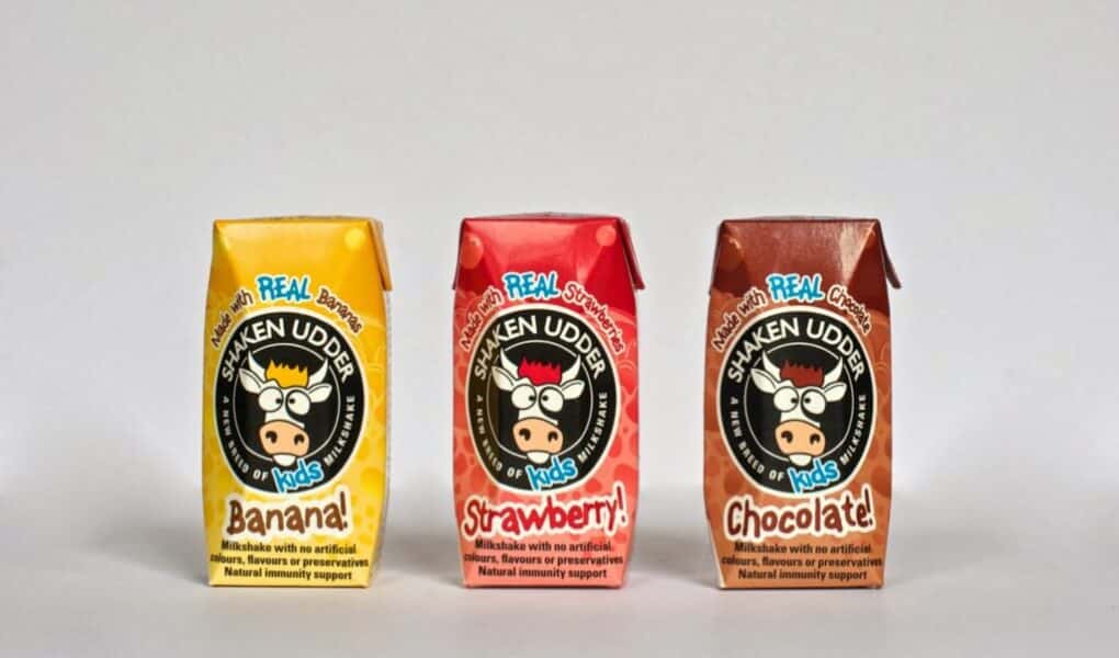 Three small, colourful cartons of Shaken Udder Kids milk drinks are arranged in a row. Flavours shown are Banana (yellow carton), Strawberry (red carton), and Chocolate (brown carton). Each features a cartoon cow logo and playful text.