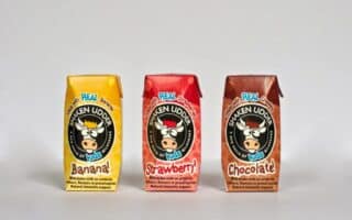 Three small, colourful cartons of Shaken Udder Kids milk drinks are arranged in a row. Flavours shown are Banana (yellow carton), Strawberry (red carton), and Chocolate (brown carton). Each features a cartoon cow logo and playful text.