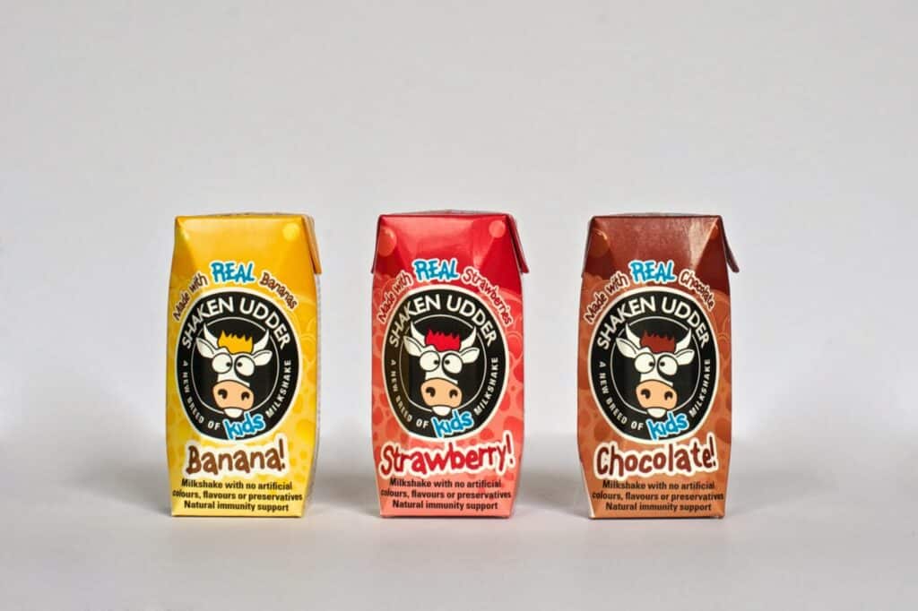 Three small Shaken Udder milkshake cartons for children are lined up side by side. Flavours are banana (yellow box), strawberry (red box), and chocolate (brown box). Each carton shows a cartoon cow face and product details on the front.
